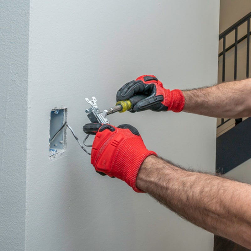 Electrician installing outlet in wall with red gloves, using screwdriver.