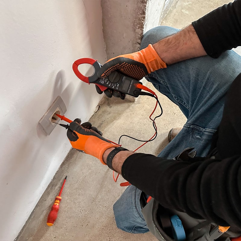 Electrician testing wall outlet voltage with a multimeter and wearing gloves for safety.