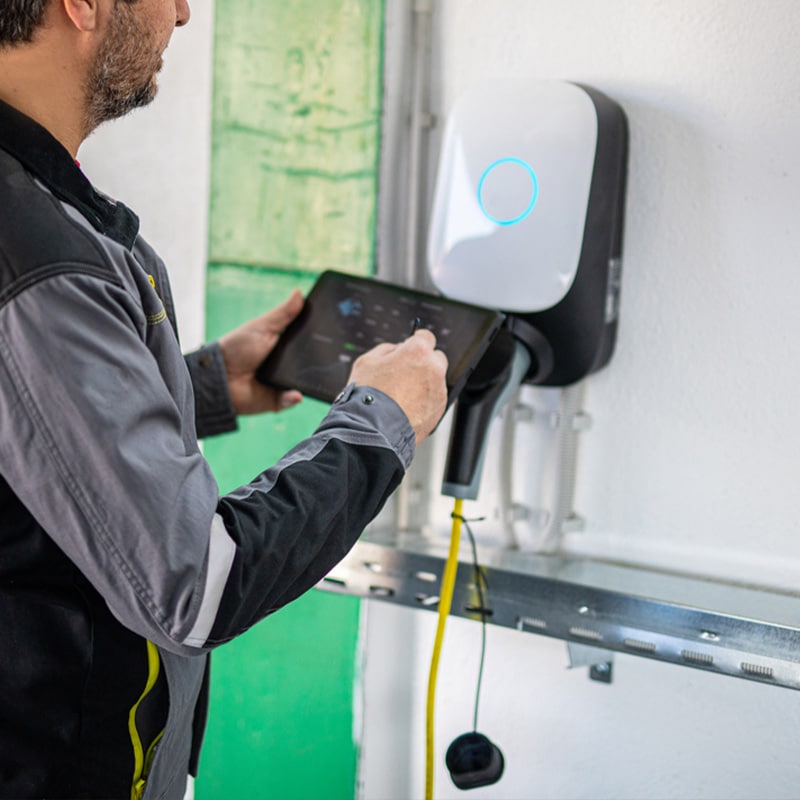 Technician using a tablet to operate a wall-mounted electric vehicle charging station indoors.