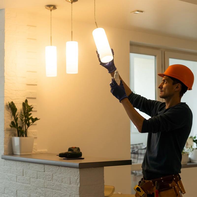 Electrician installing hanging lights in modern kitchen, wearing hard hat and gloves. Home lighting upgrade in progress.