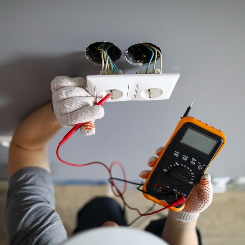 Electrician testing wall socket voltage with multimeter, wearing protective gloves for safety.