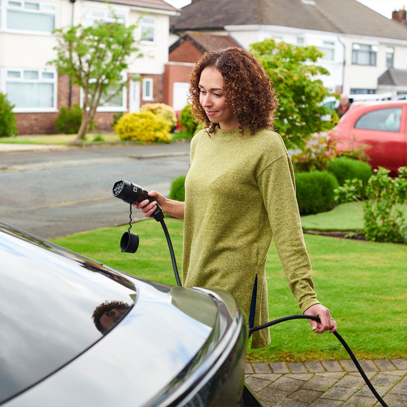 Woman charging electric car at home in a residential neighborhood on a sunny day.