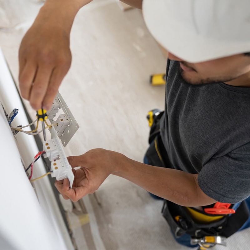 Electrician in a hard hat installing a wall outlet with tools, ensuring safe wiring connections.
