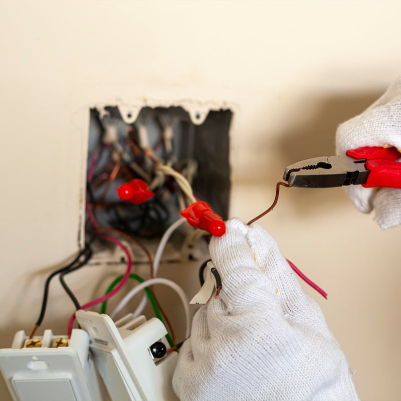 Electrician using pliers to work on electrical wires in a wall outlet box, wearing protective gloves.