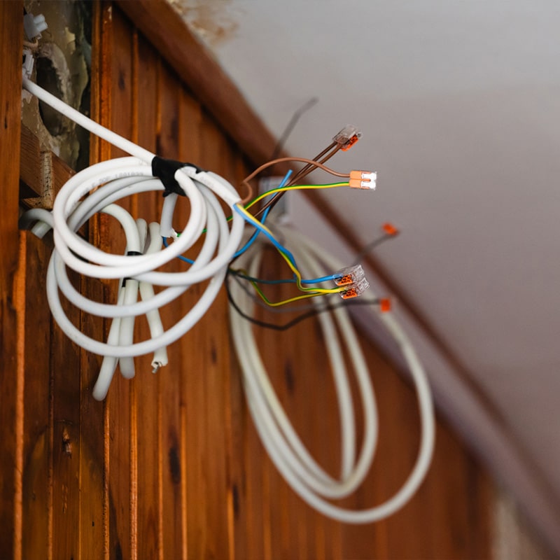 Exposed electrical wiring hanging from wooden wall corner, showing home renovation or repair work in progress.