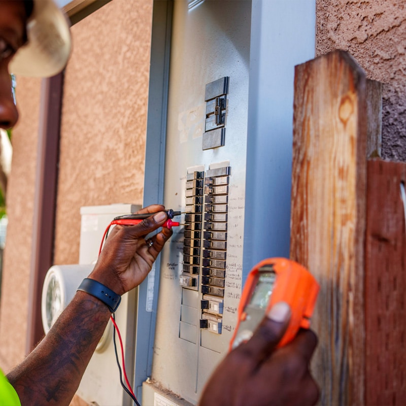 Electrician inspecting and testing electrical panel with a multimeter outdoors.