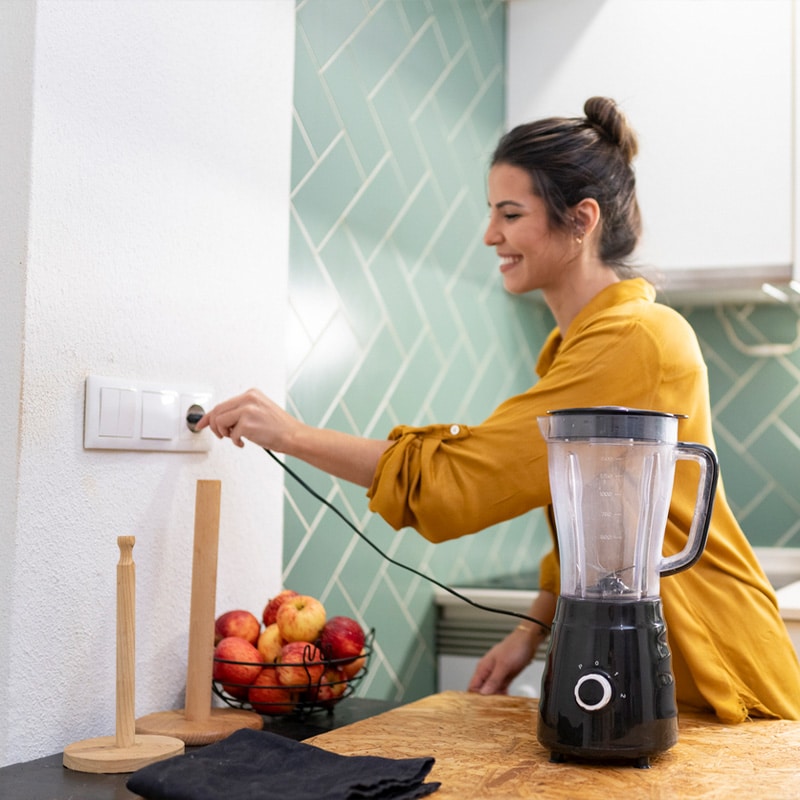 Woman in yellow shirt plugging in a blender in kitchen with green tiles and apples on counter.