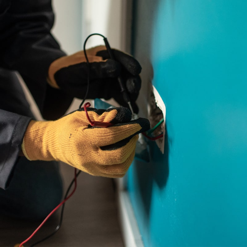Electrician wearing gloves works on wiring in wall socket, using a tester tool for safe electrical repair.