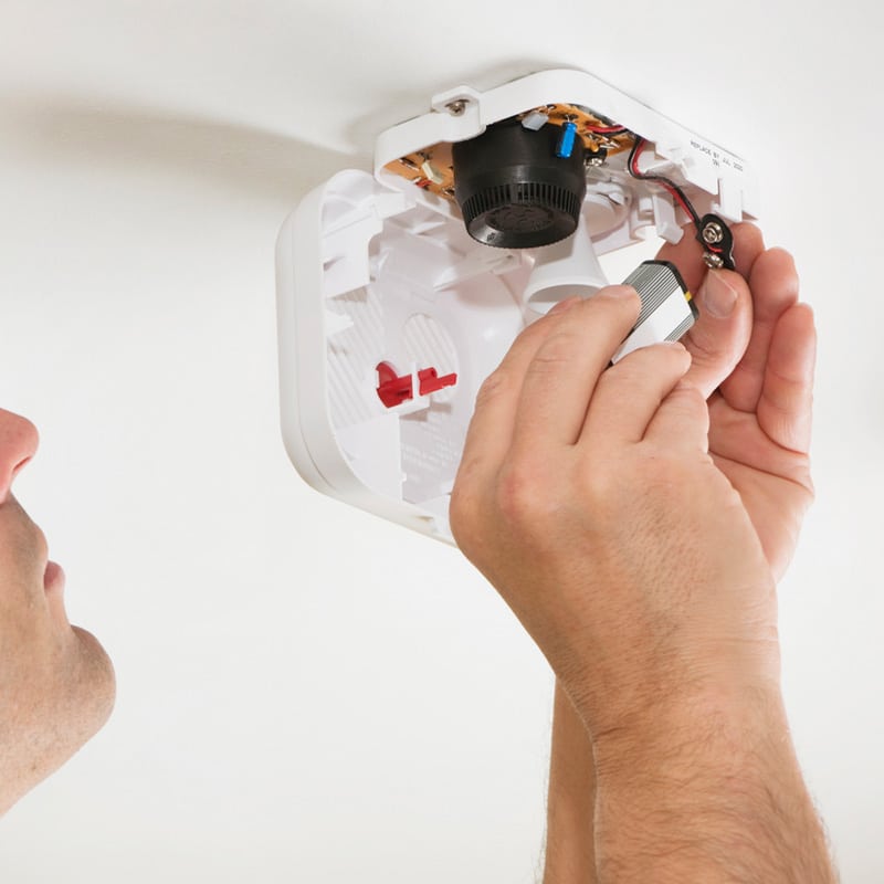 Person replacing battery in smoke detector on ceiling for maintenance and safety.